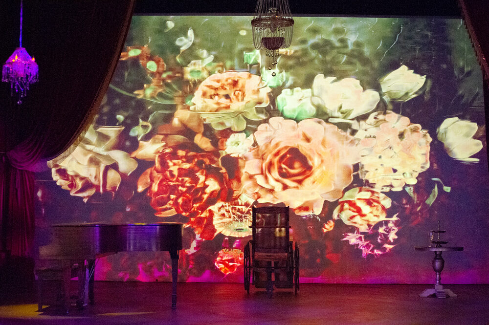 A stage dressed with a painted floral projection, chandelier and crimson drape