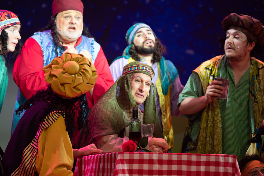 An ensemble in bright character costume around a table mid-scene