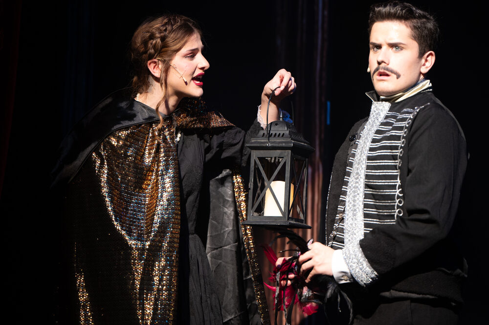 Two performers close together on a darkened stage, one holding a lantern